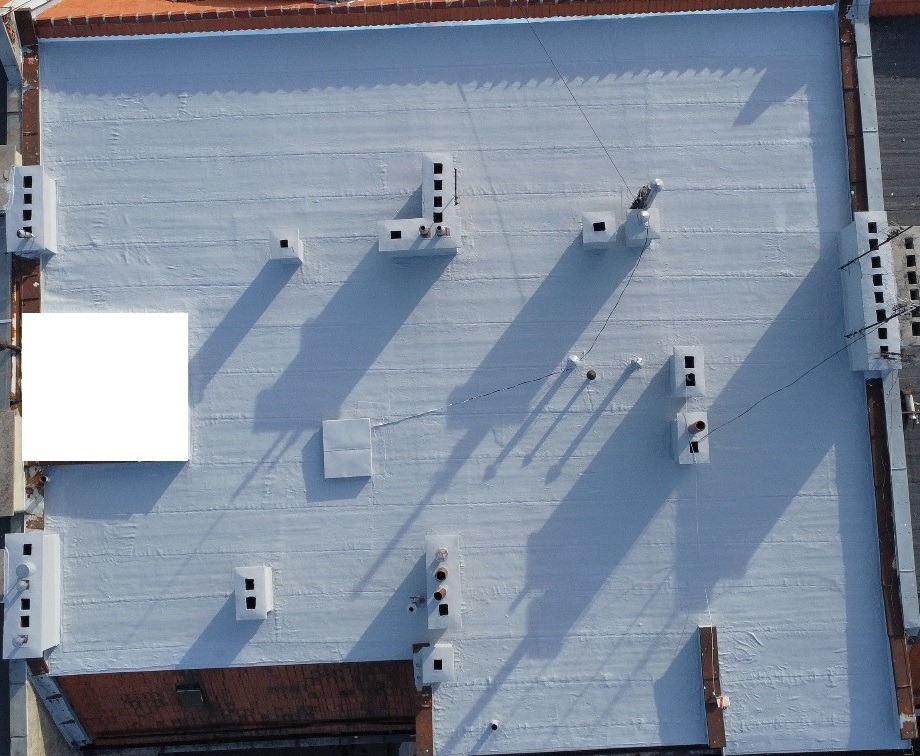 Widok z góry na płaski, pomalowany na szaro dach budynku z licznymi kominami wentylacyjnymi i instalacjami, z widocznymi śladami po naprawach i uszczelnieniach.