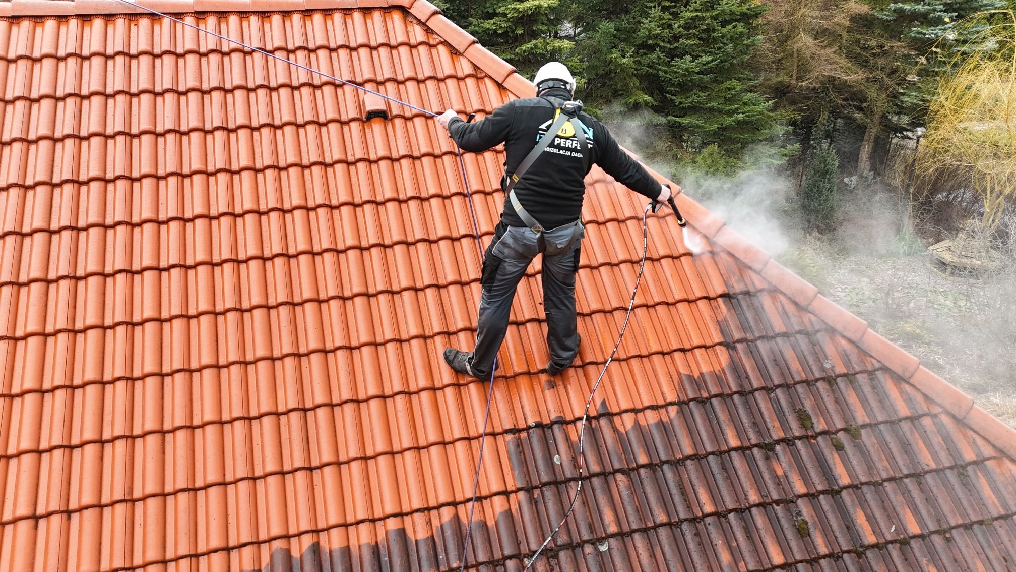 Pracownik w uprzęży czyści dachówkę ciśnieniowo. Część dachu umyta, widoczna różnica w kolorze. Tło stanowią drzewa.