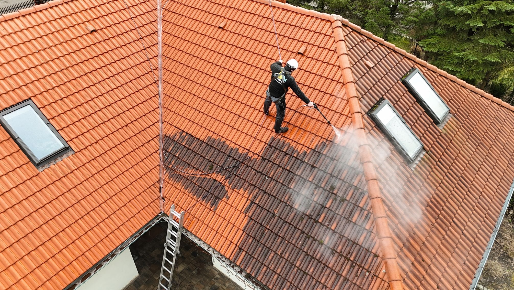 Czerwona dachówka czyszczona myjką ciśnieniową przez pracownika w uprzęży, widok z góry. Część dachu umyta, część brudna. Obok drabina i okna dachowe.