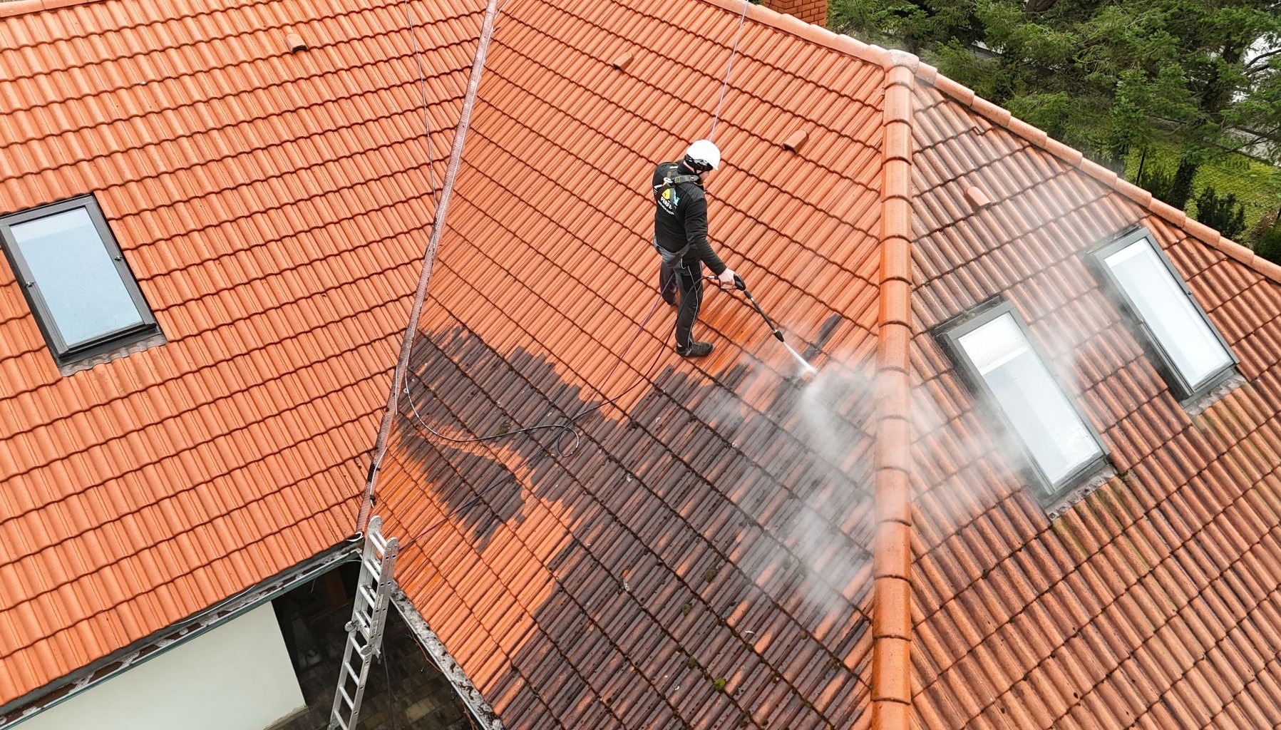 Ceglasty dach z oknami połaciowymi, częściowo umyty myjką ciśnieniową przez pracownika w kasku. Widoczna drabina i zieleń w tle. Ujęcie z góry.