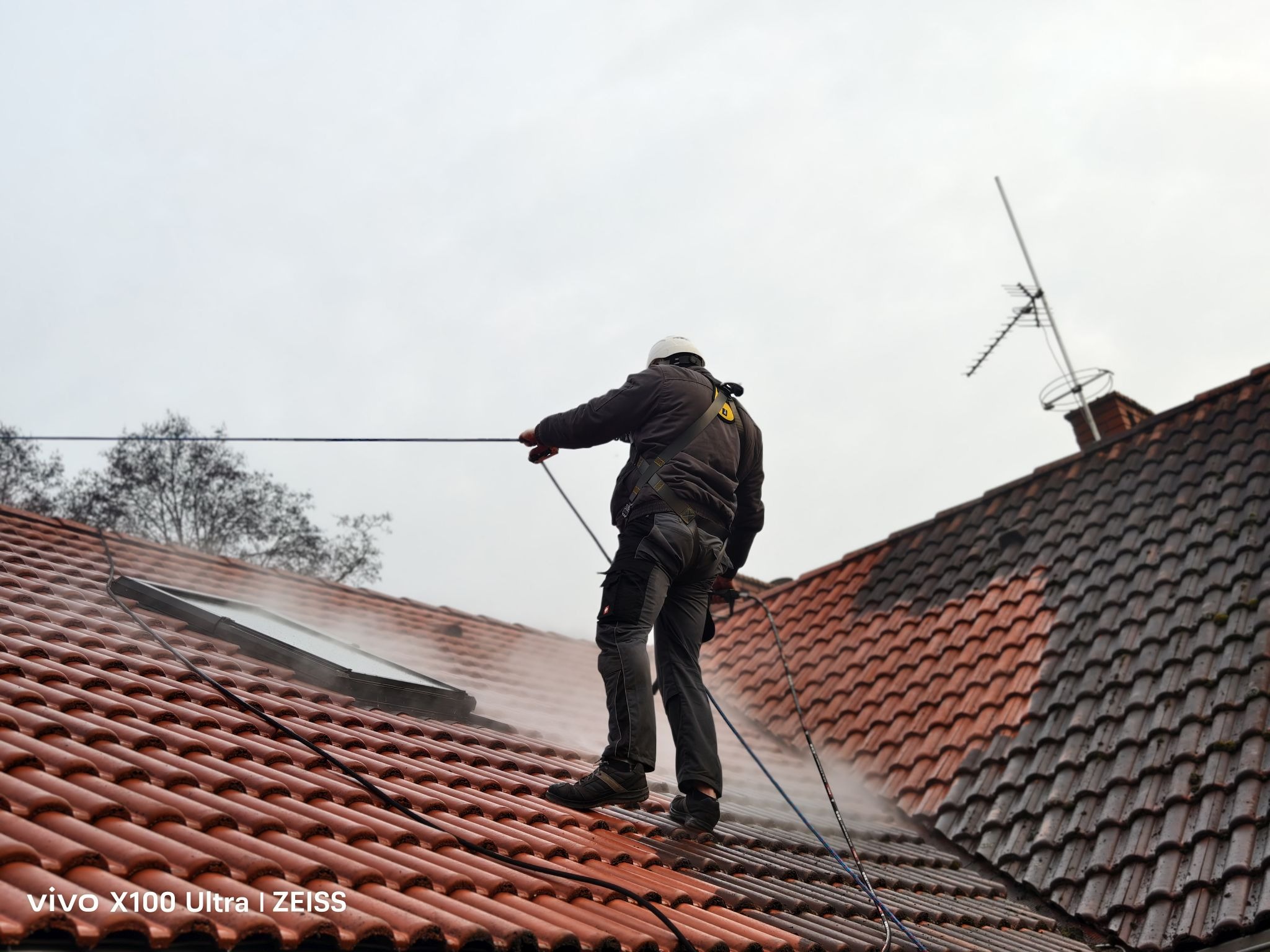 Pracownik w uprzęży czyści dachówkę pod ciśnieniem, widoczna różnica w czystości połaci. Antena na kominie w tle, pochmurne niebo.