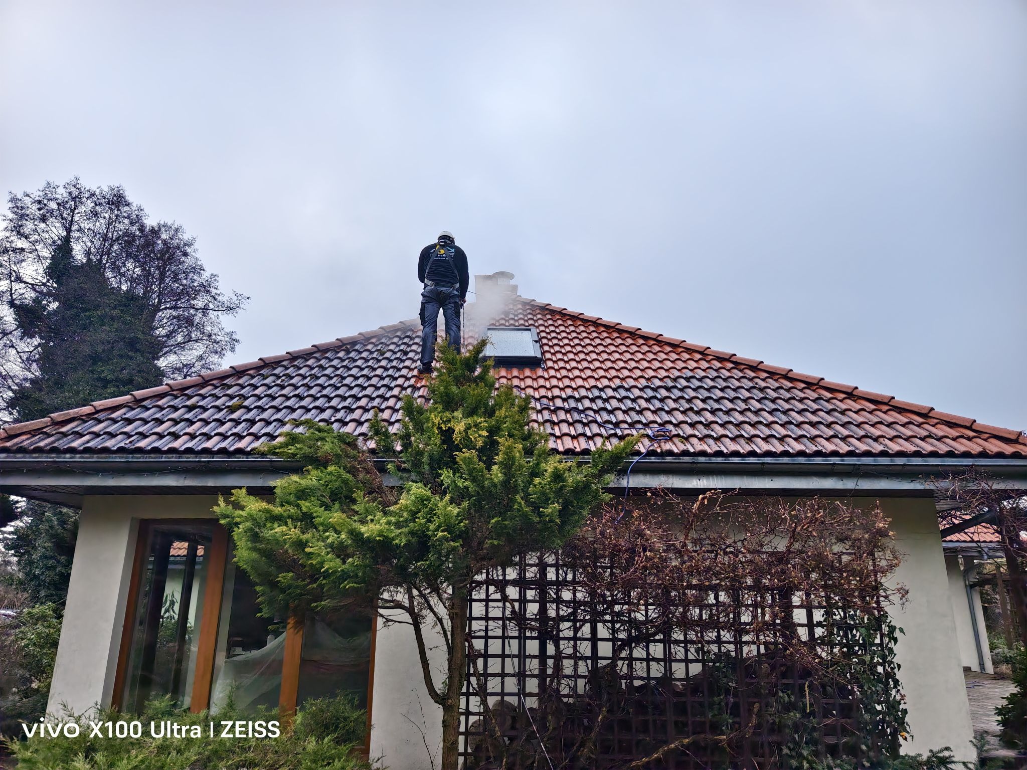 Pracownik w kombinezonie roboczym czyści dach z czerwonej dachówki za pomocą myjki ciśnieniowej, stojąc na szczycie dachu. Widoczny budynek i zieleń.
