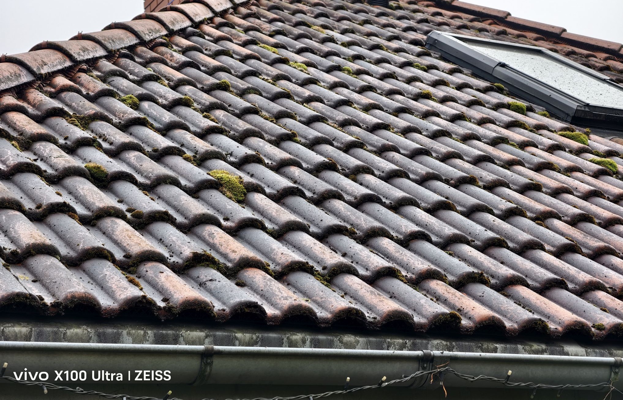 Czerwona dachówka pokryta mchem, widok z bliska. W tle okno dachowe i fragment rynny. Widoczne zabrudzenia i ślady wilgoci na powierzchni.