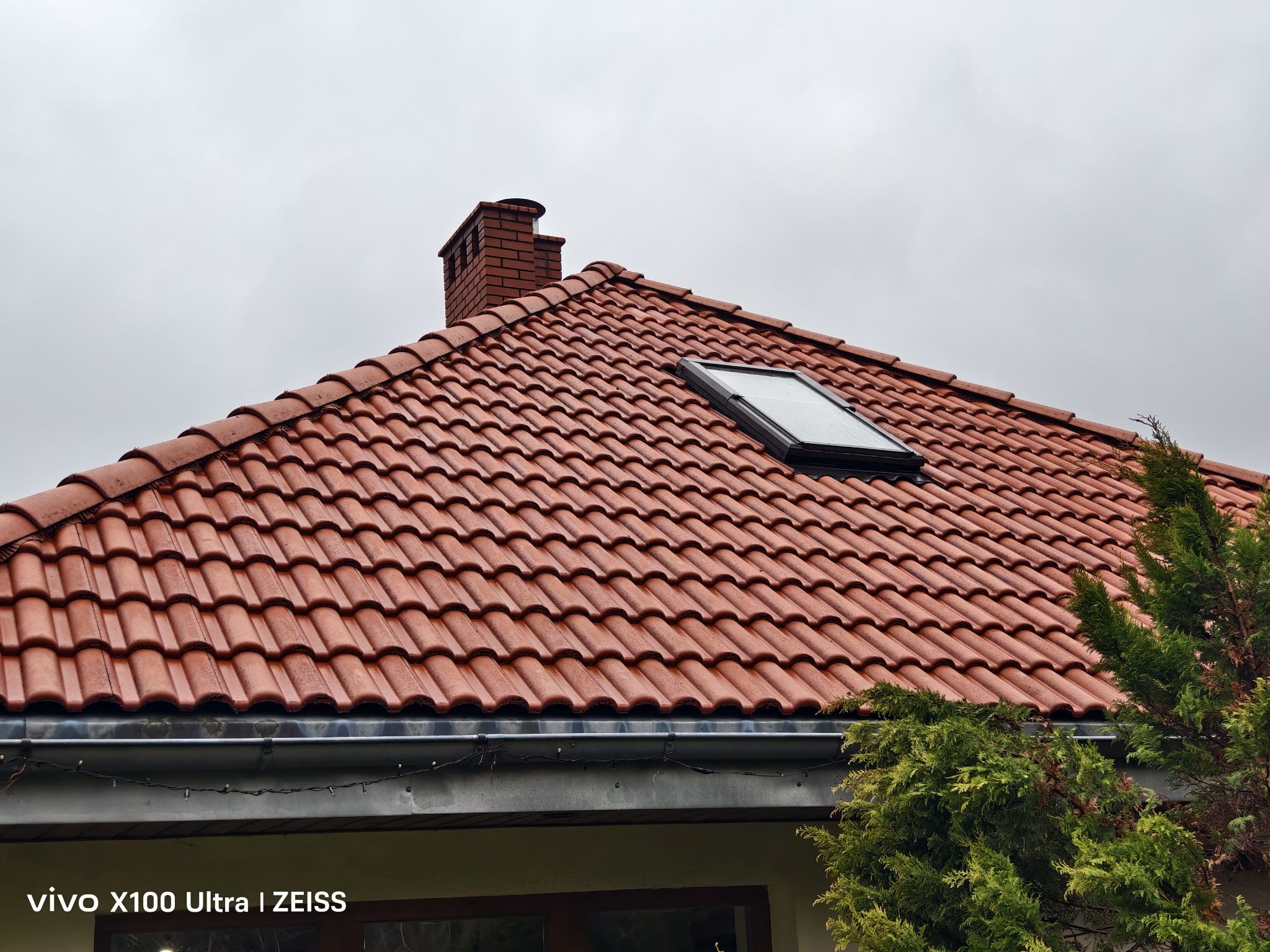 Czerwona dachówka ceramiczna z oknem dachowym i kominem z cegły na tle pochmurnego nieba. Widoczne rynny i fragment zieleni.