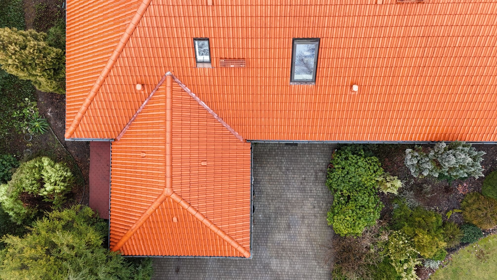 Widok z góry na dach z czerwonej dachówki ceramicznej z dwoma oknami połaciowymi, otoczony zielenią.