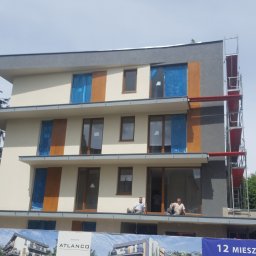 ANWIL KRZYSZTOF ŁAJSZCZAK - Budynek wielorodzinny z rusztowaniem i oknami zabezpieczonymi folią. Dwóch pracowników siedzi na wystającym elemencie elewacji.