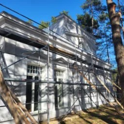 Elewacja budynku w stylu klasycystycznym, z detalami architektonicznymi, otoczona rusztowaniem i drzewami w słoneczny dzień. Widoczne elementy dekoracyjne.