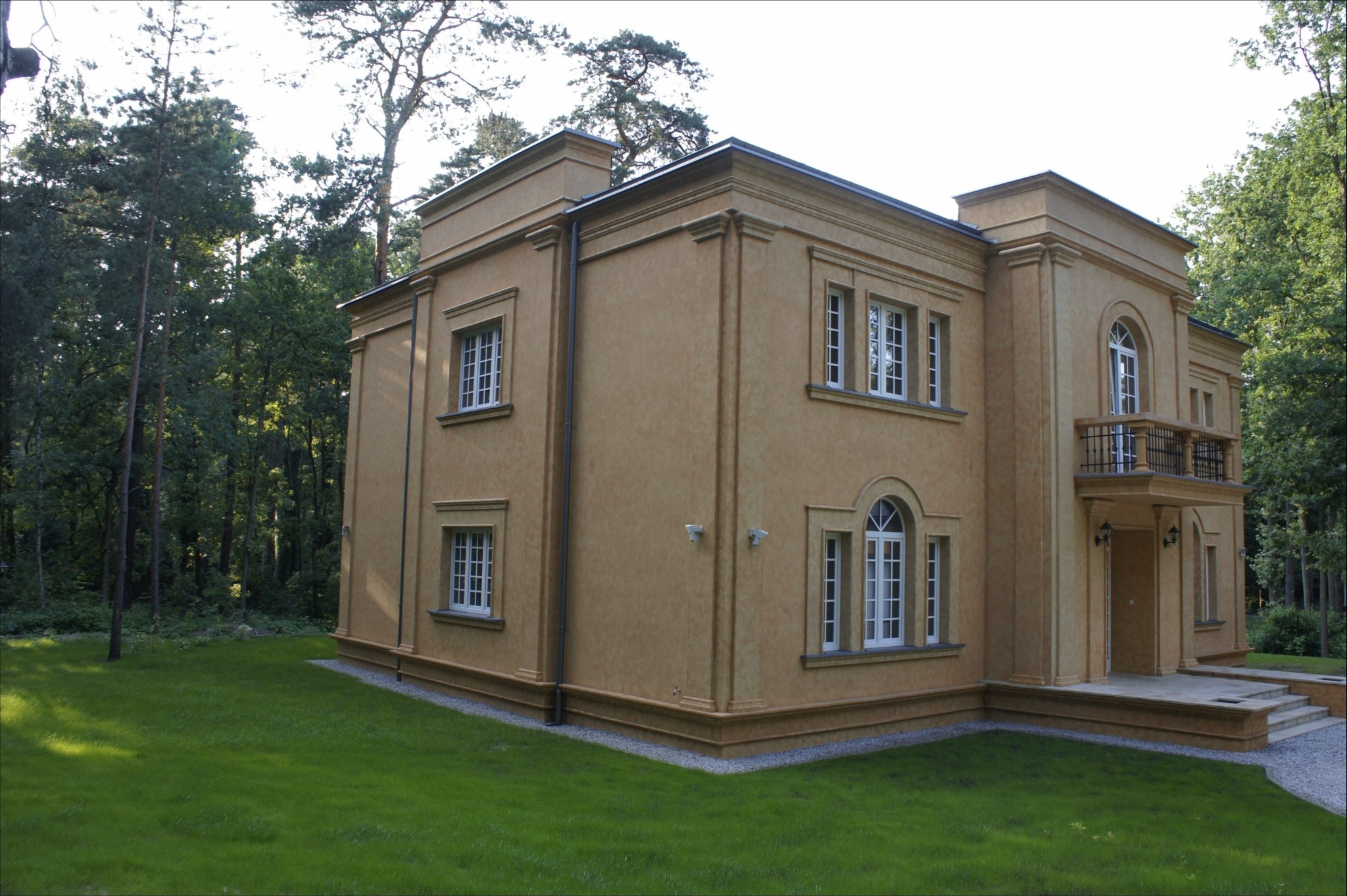 Elegancki, dwukondygnacyjny dom z beżową elewacją i balkonem, otoczony zielonym trawnikiem i drzewami. Widoczne detale architektoniczne i oświetlenie zewnętrzne.