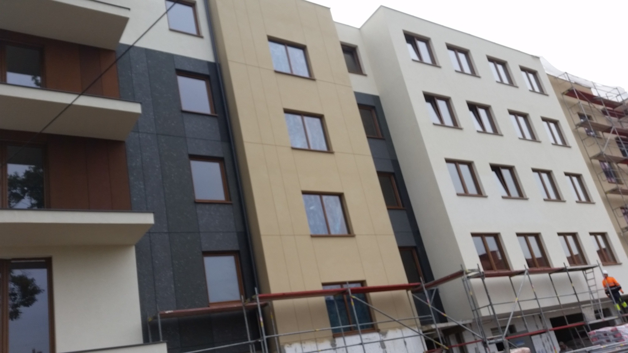 Nowoczesny budynek wielorodzinny z elewacją w odcieniach bieli, beżu i grafitu. Widoczne rusztowania i pracownik w pomarańczowym kombinezonie.