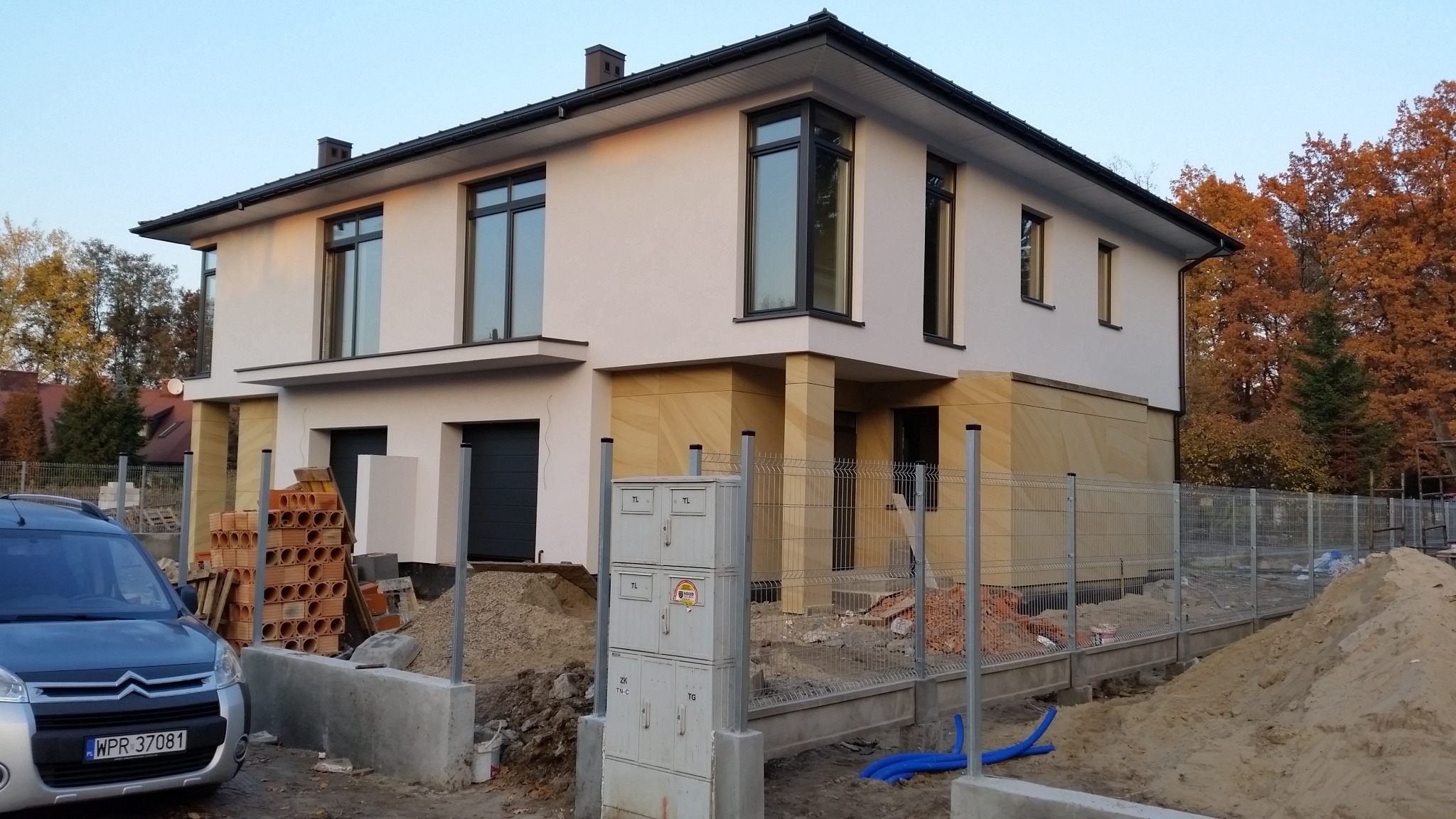 Dwukondygnacyjny dom z elewacją w odcieniach beżu i bieli, z widocznymi elementami wykończeniowymi i materiałami budowlanymi na posesji.