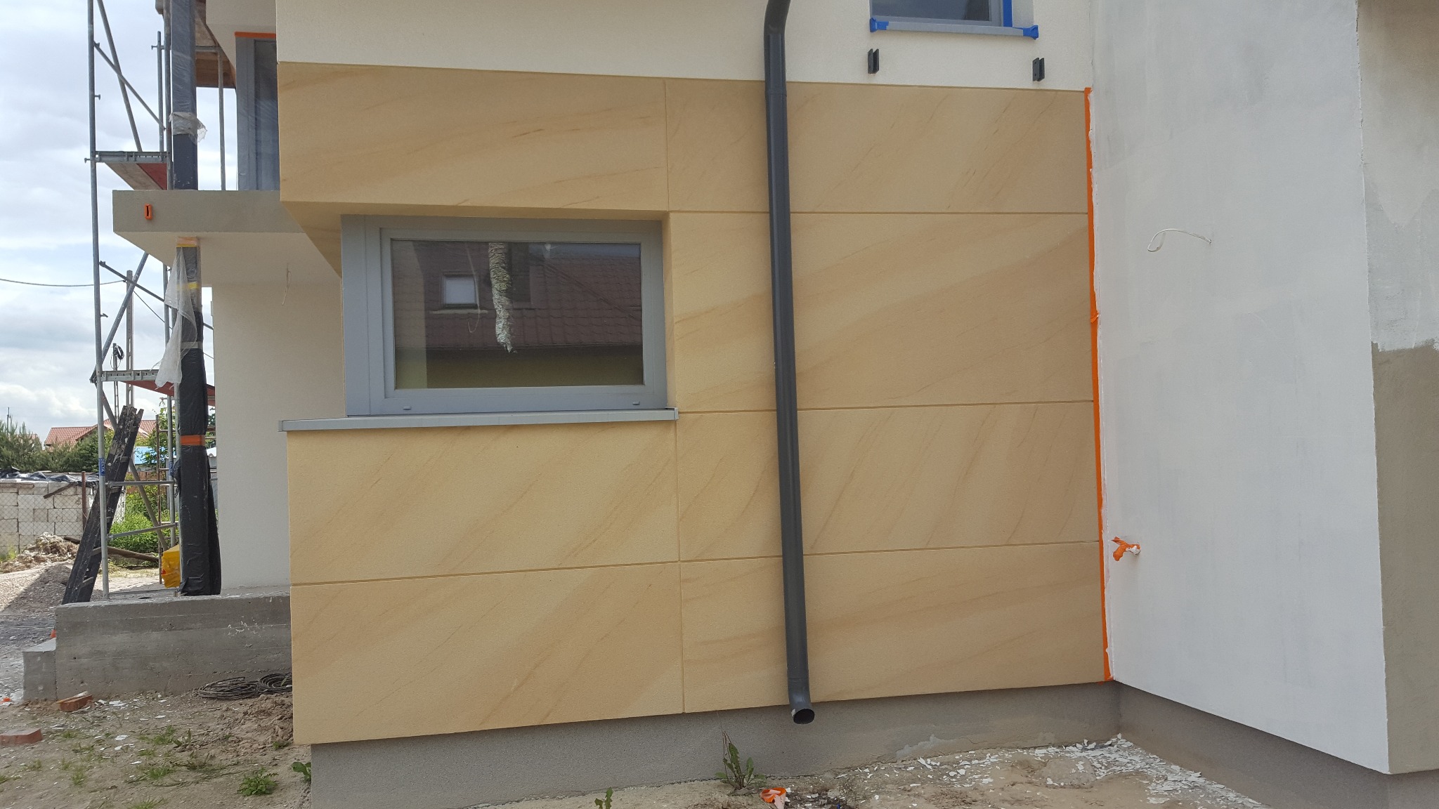 Elewacja domu z panelami piaskowymi, szara rynna i okno. Część ściany pomalowana na biało. Widoczne rusztowanie i teren budowy.