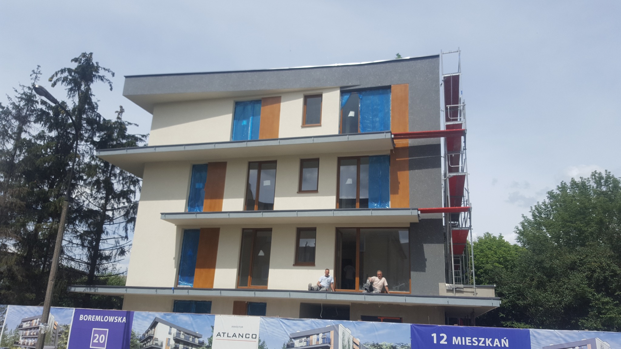 Budynek wielorodzinny z rusztowaniem i oknami zabezpieczonymi folią. Dwóch pracowników siedzi na wystającym elemencie elewacji.