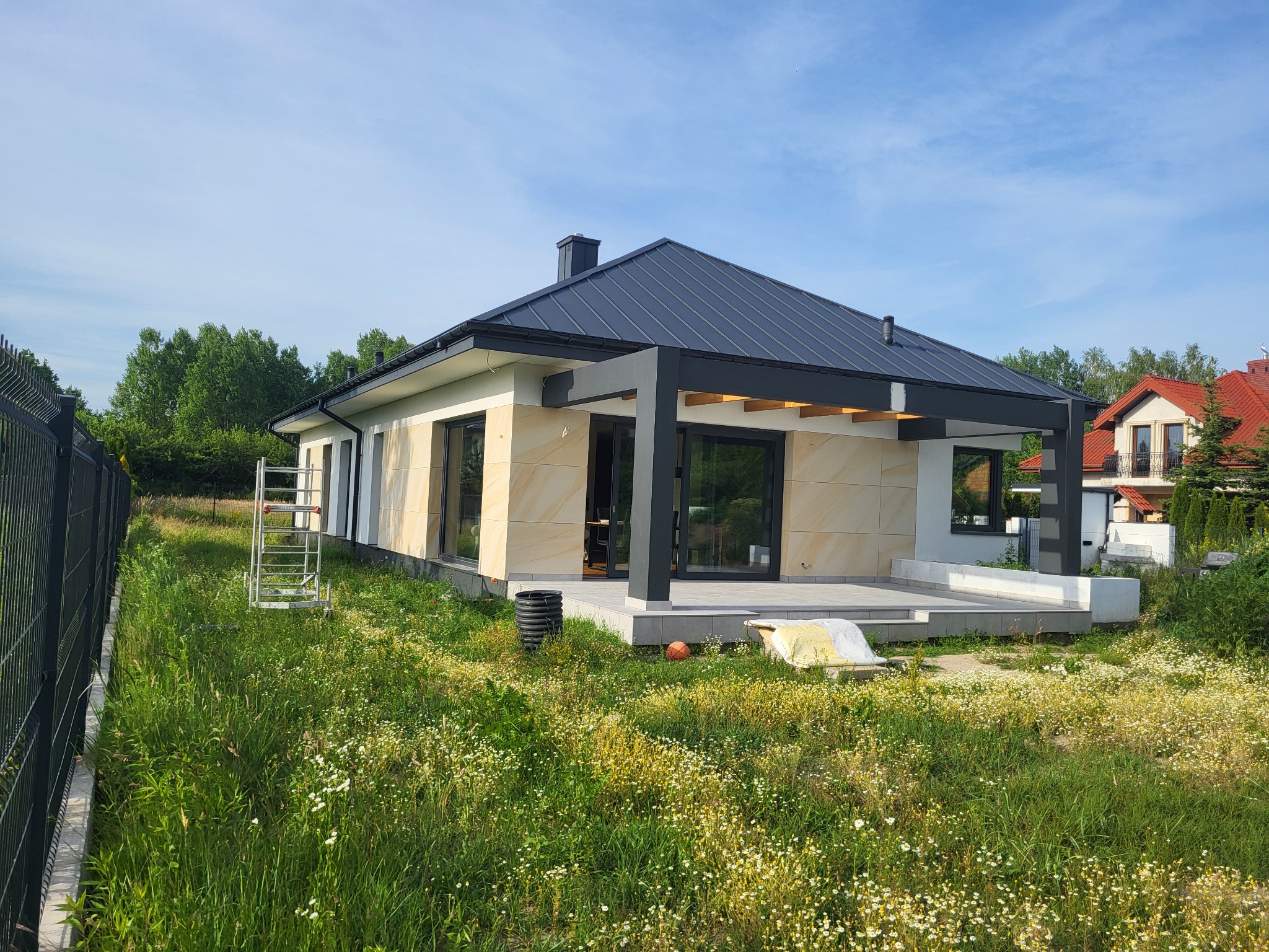 Nowoczesny dom jednorodzinny z elewacją z jasnego kamienia i antracytowym dachem, widok z ogrodu z bujną roślinnością i metalowym ogrodzeniem.