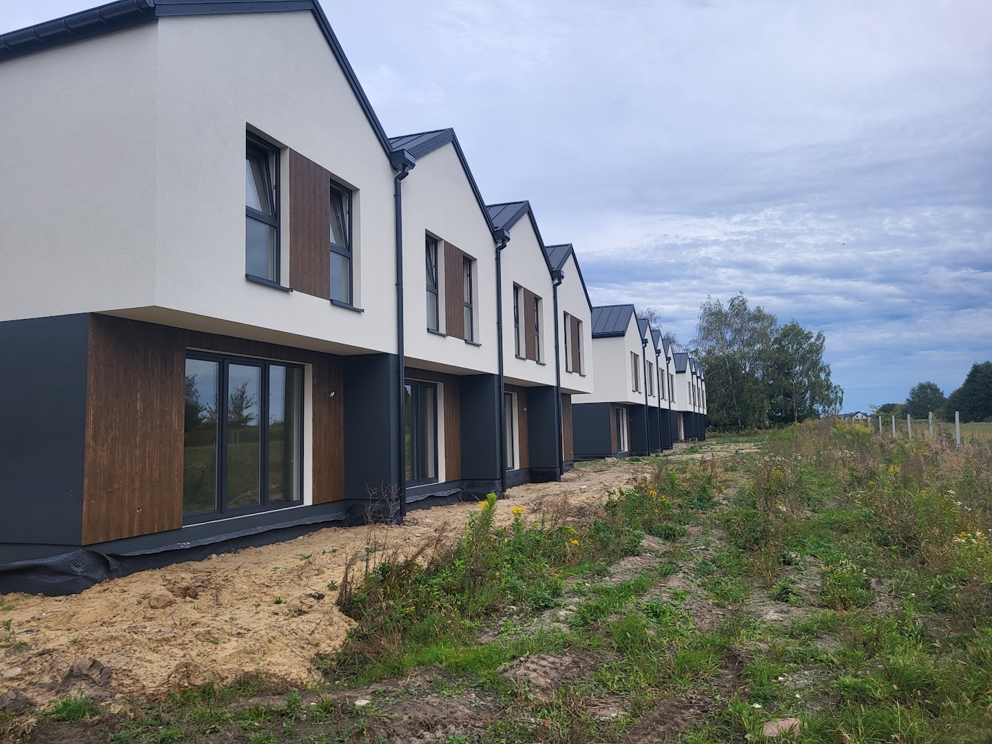 Szereg nowoczesnych domów z białą elewacją i drewnianymi panelami, widoczne z zewnątrz, w surowym otoczeniu. Domy mają czarne dachy i okna.