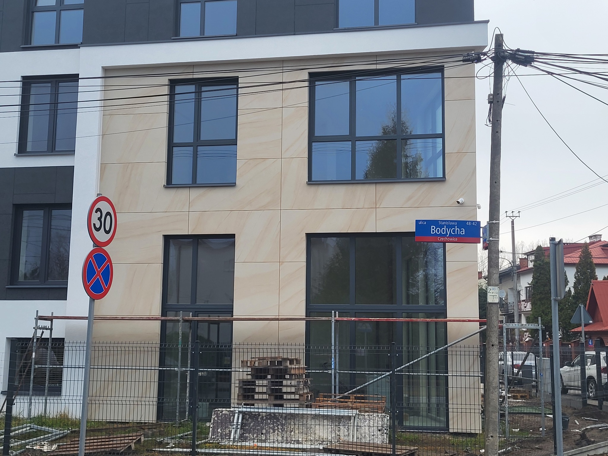 Nowoczesna elewacja budynku z beżowymi płytami i ciemnymi oknami, widoczny znak drogowy i tabliczka z nazwą ulicy. Na pierwszym planie ogrodzenie budowlane.