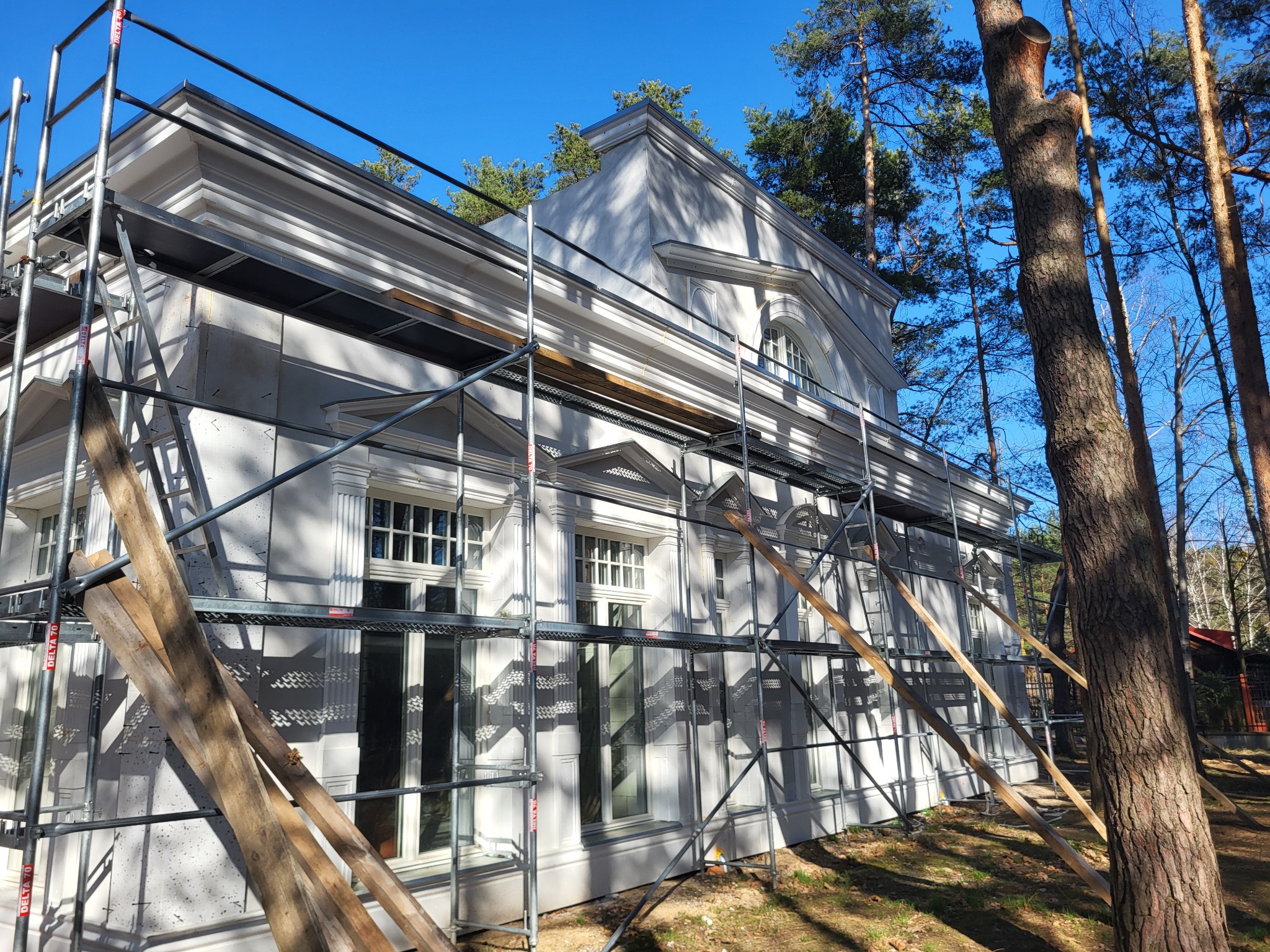 Elewacja budynku w stylu klasycystycznym, z detalami architektonicznymi, otoczona rusztowaniem i drzewami w słoneczny dzień. Widoczne elementy dekoracyjne.