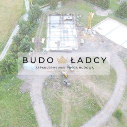 Budowładcy sp. z o.o. - Widok z drona na plac budowy z przygotowanymi fundamentami, szalunkami i zbrojeniami, widoczny również żuraw budowlany oraz logo firmy Budowładcy.