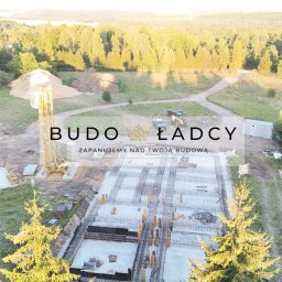 Budowładcy sp. z o.o. - Widok z lotu ptaka na plac budowy z widocznymi fundamentami, zbrojeniami, żurawiem, stertą ziemi i okolicznymi drzewami; w tle las i jezioro. Na pierwszym planie logo firmy budowlanej.