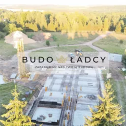 Widok z lotu ptaka na plac budowy z widocznymi fundamentami, zbrojeniami, żurawiem, stertą ziemi i okolicznymi drzewami; w tle las i jezioro. Na pierwszym planie logo firmy budowlanej.