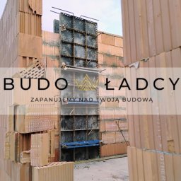 Budowładcy sp. z o.o. - Wznoszenie ściany z bloczków ceramicznych Porotherm, widoczne szalunki do wylewania słupów żelbetowych, pręty zbrojeniowe na pierwszym planie, logo firmy nałożone na zdjęcie.