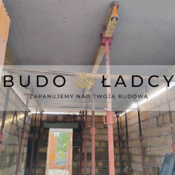 Budowładcy sp. z o.o. - Wnętrze budynku w trakcie budowy, ściany z cegły ceramicznej, betonowy strop podparty stemplami budowlanymi, widok na zewnątrz przez otwór drzwiowy.
