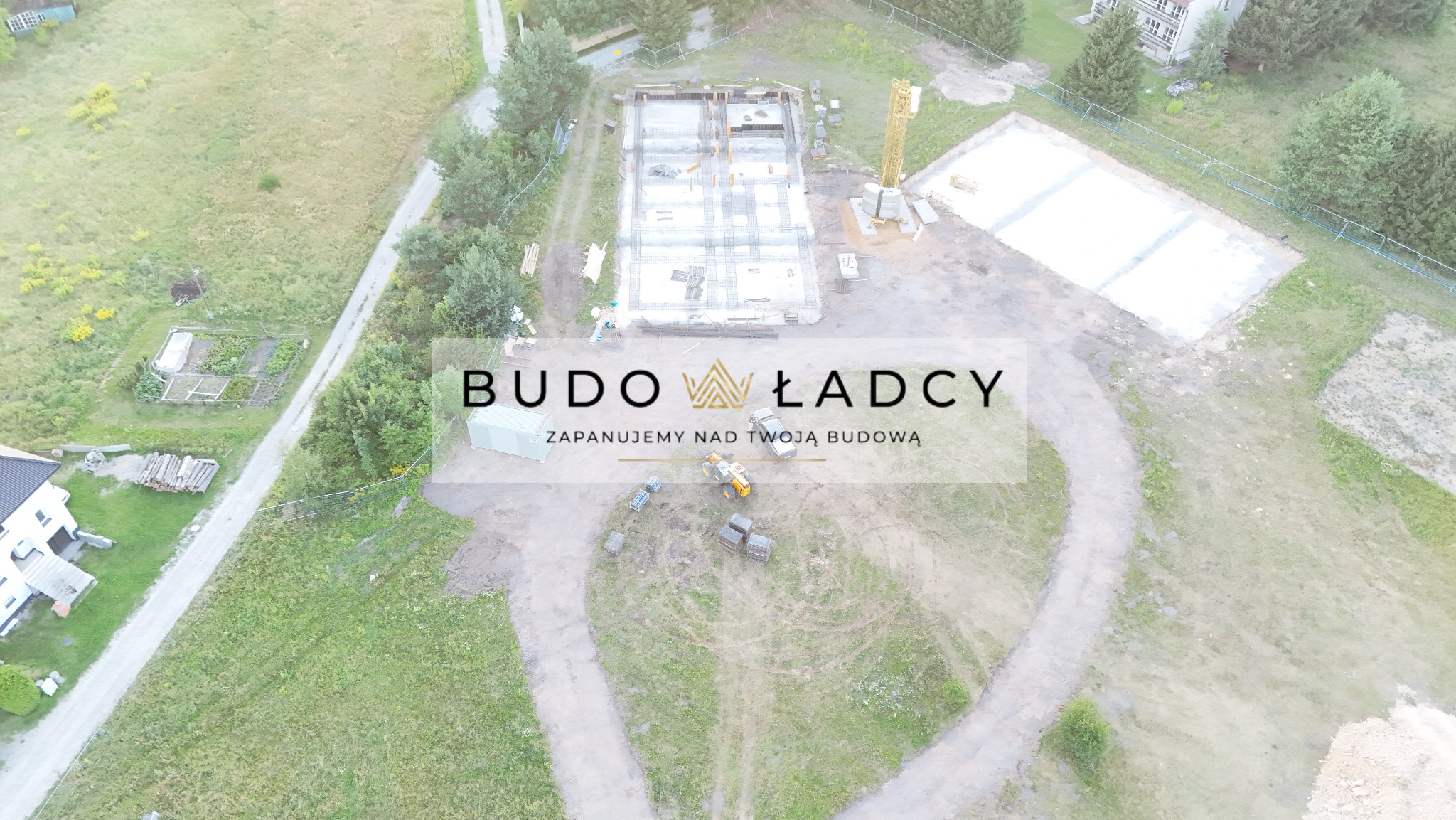 Widok z drona na plac budowy z przygotowanymi fundamentami, szalunkami i zbrojeniami, widoczny również żuraw budowlany oraz logo firmy Budowładcy.
