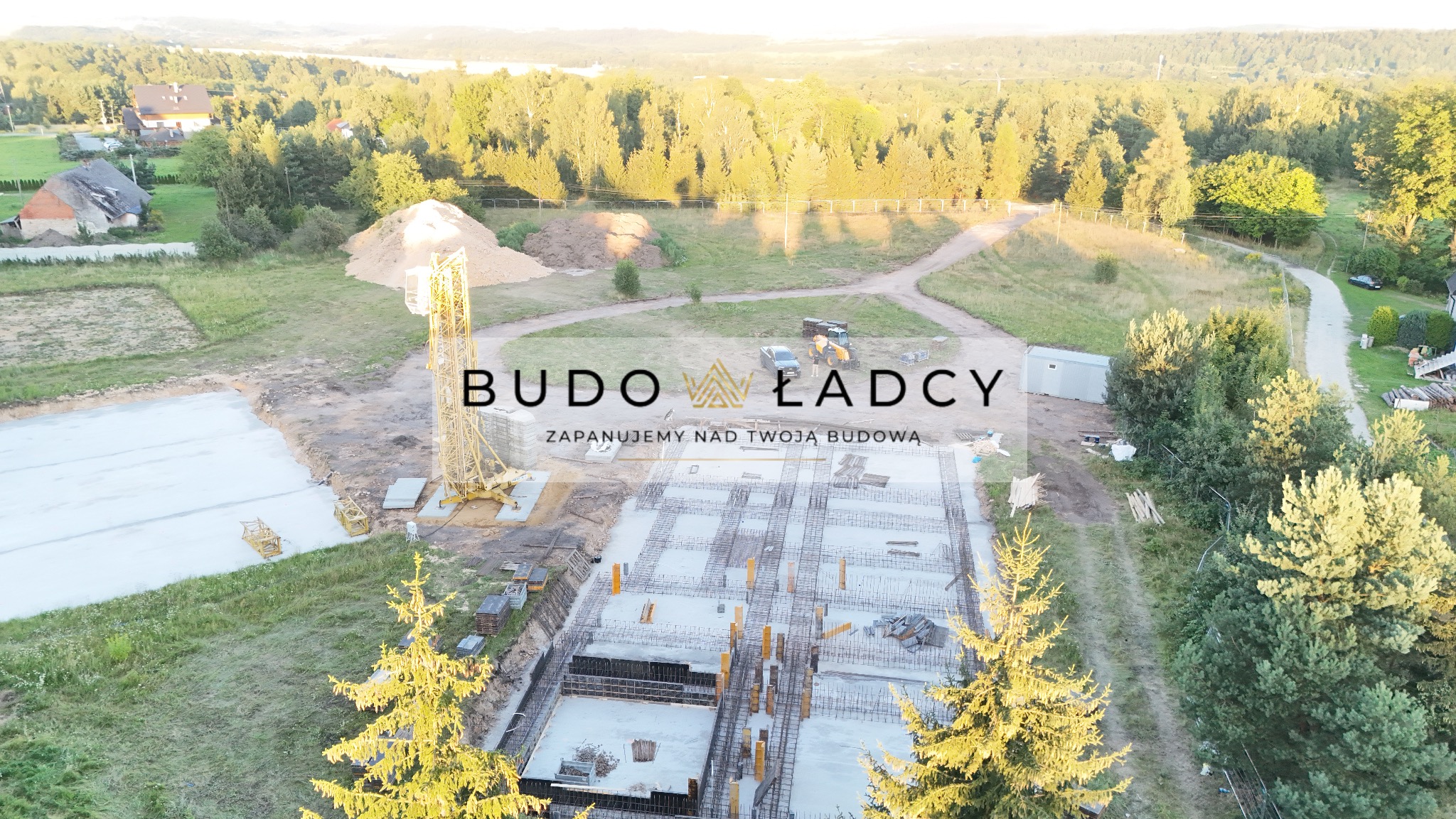 Widok z lotu ptaka na plac budowy z widocznymi fundamentami, zbrojeniami, żurawiem, stertą ziemi i okolicznymi drzewami; w tle las i jezioro. Na pierwszym planie logo firmy budowlanej.