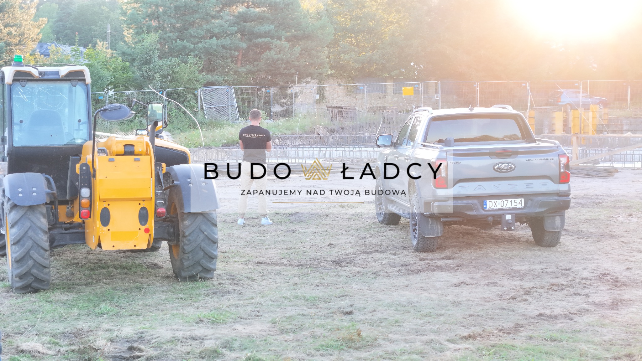 Żółta ładowarka teleskopowa i szary pickup Ford Ranger na placu budowy z widocznymi zbrojeniami fundamentów w tle, osoba w koszulce z logo firmy budowlanej stoi obok.