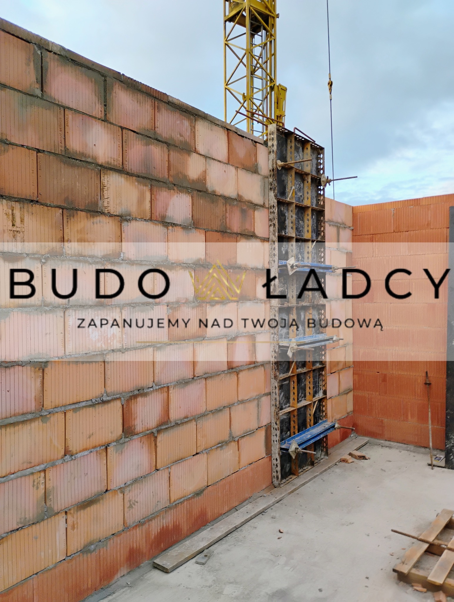 Wznoszenie ściany z czerwonych bloczków ceramicznych z widocznym systemem szalunkowym i żółtym dźwigiem w tle, na placu budowy.