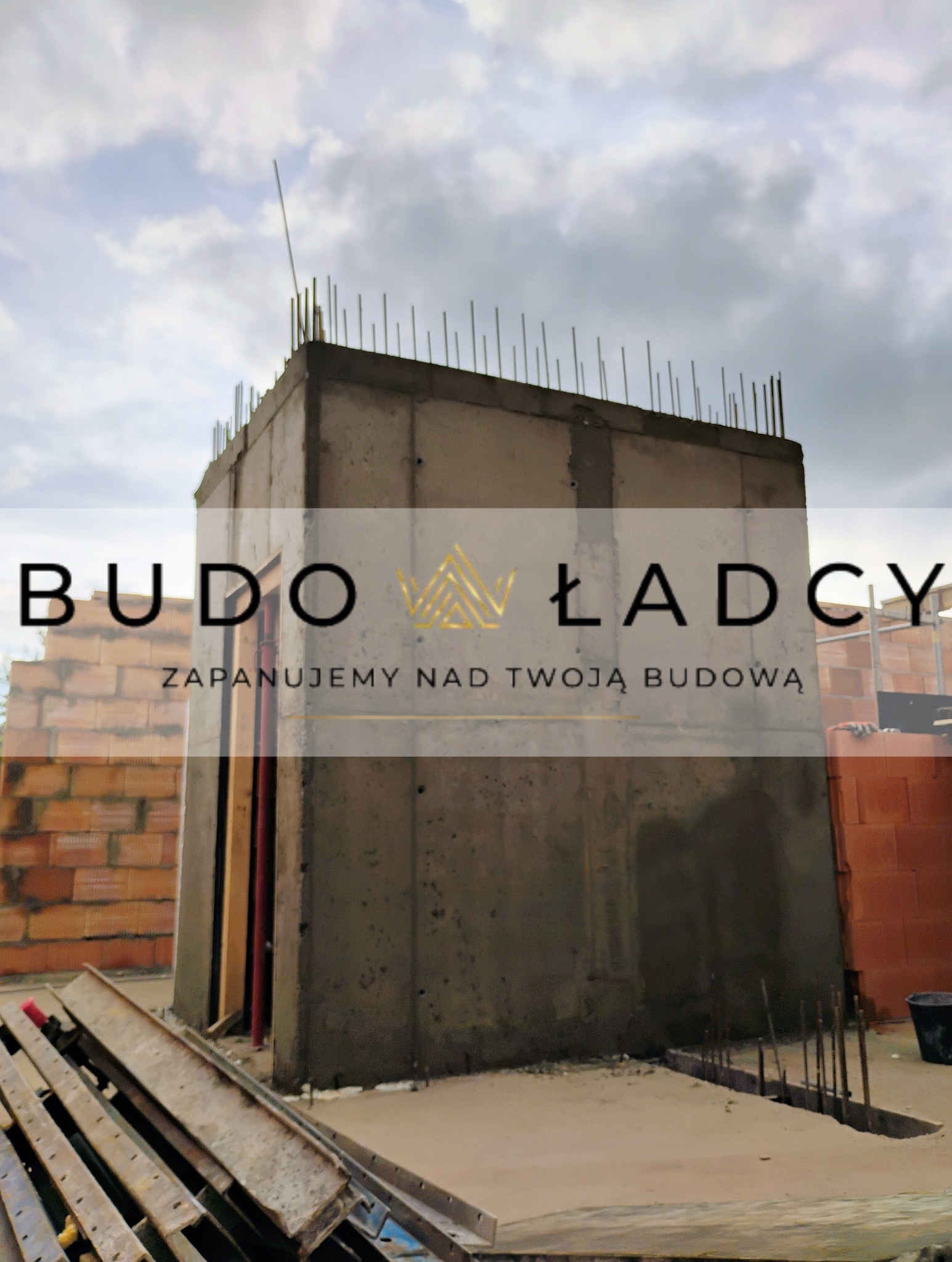 Żelbetowy szyb windowy w trakcie budowy, wystające pręty zbrojeniowe, częściowo widoczne ściany z pustaków, szare niebo w tle.