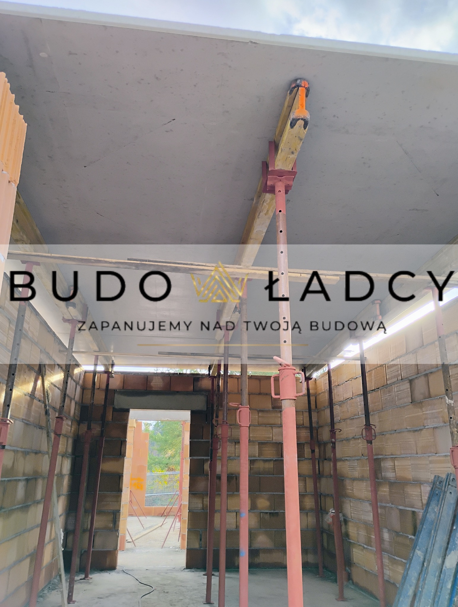 Wnętrze budynku w trakcie budowy, ściany z cegły ceramicznej, betonowy strop podparty stemplami budowlanymi, widok na zewnątrz przez otwór drzwiowy.