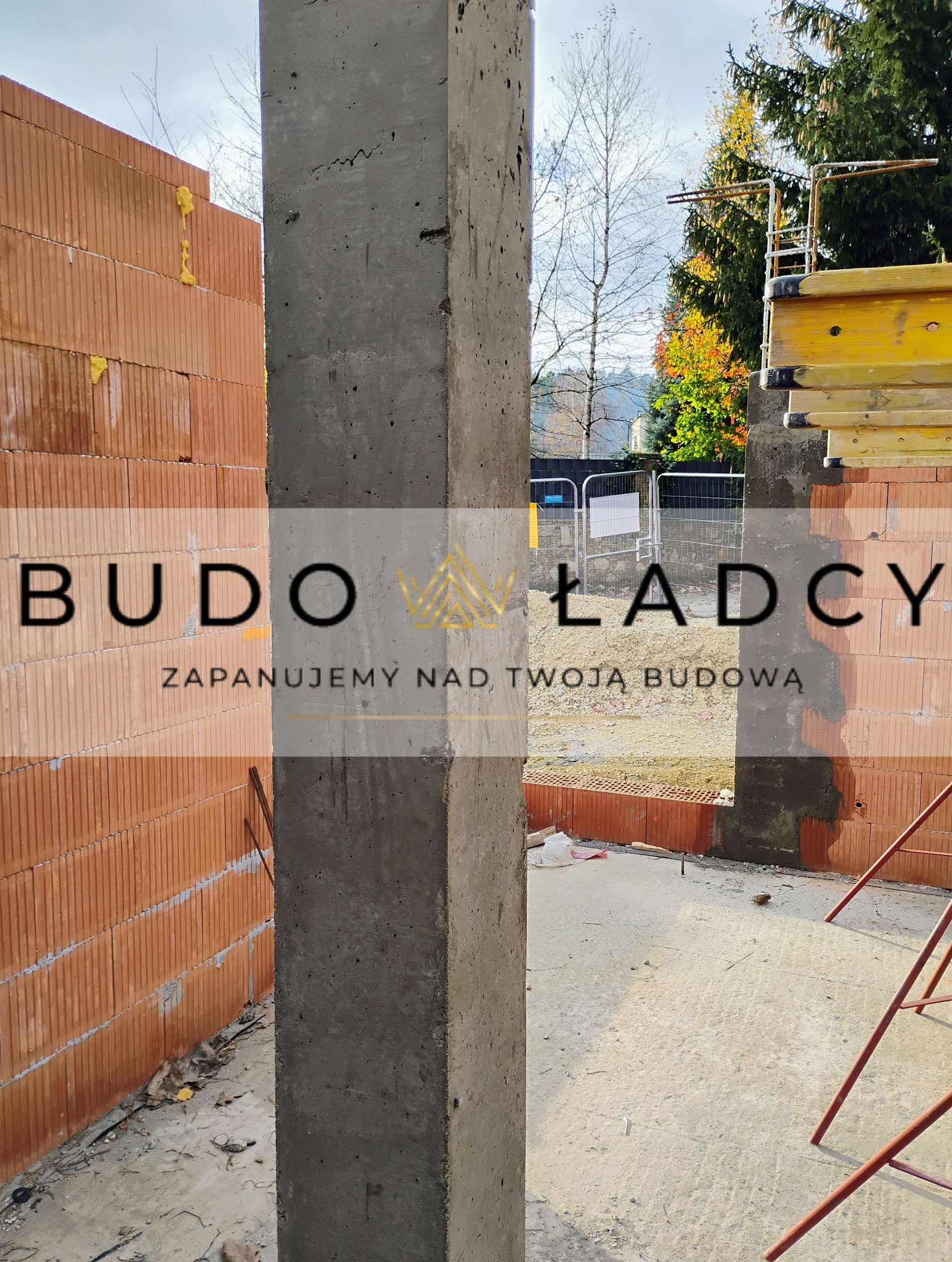 Żelbetowy słup na placu budowy, widoczne ceglane ściany w tle i elementy szalunków.