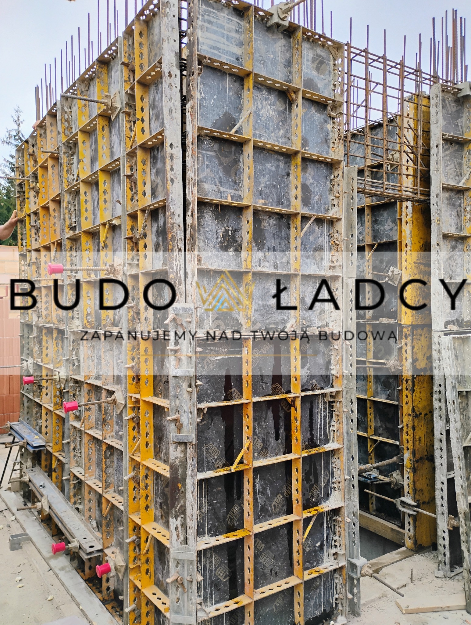 Konstrukcja szalunkowa z żółtymi elementami wzmacniającymi, wypełniona czarną membraną, przygotowana do zalewania betonem, z wystającymi prętami zbrojeniowymi.
