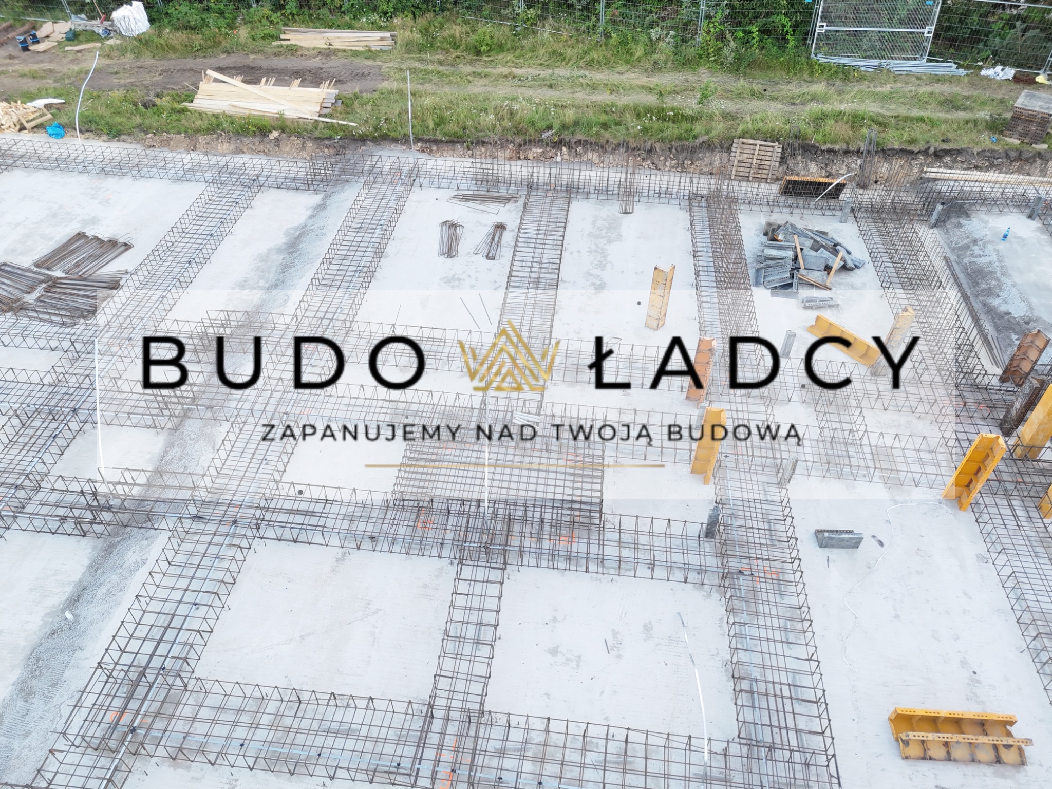 Zbrojenie fundamentów w wykopie, widok z góry, częściowo zalane betonem, z widocznymi deskowaniami i elementami konstrukcyjnymi, na zdjęciu widoczne logo firmy.