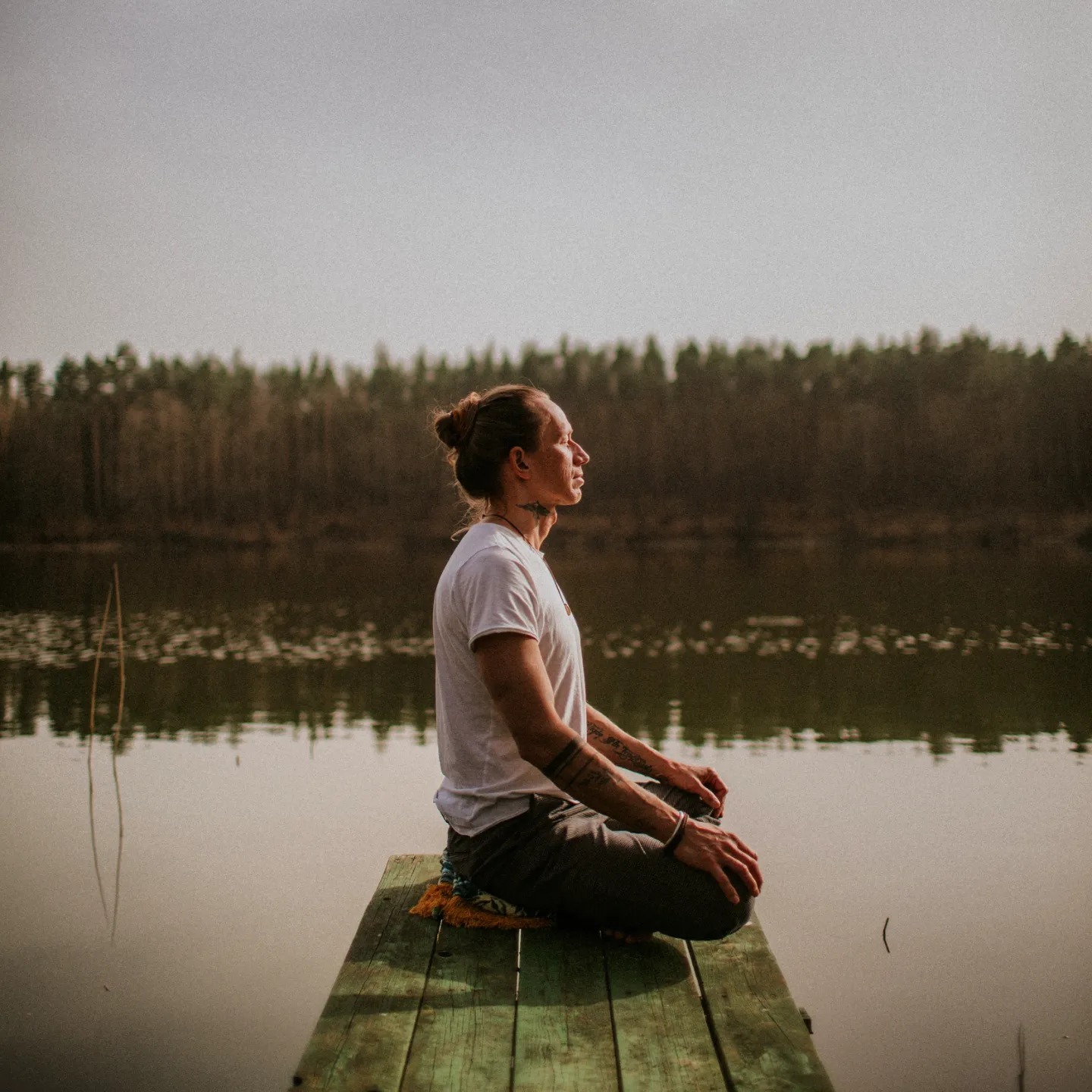Mężczyzna z tatuażami na przedramionach medytuje w pozycji lotosu na drewnianym pomoście nad spokojnym jeziorem, las w tle.