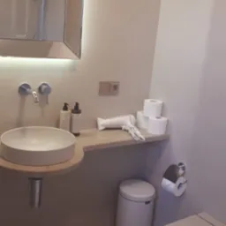 Nowoczesna łazienka z okrągłą umywalką nablatową, podświetlanym lustrem, białym koszem na śmieci i suszarką do włosów na półce, minimalistyczny design.