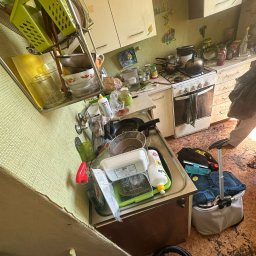 Ład @komfort - Zaniedbana kuchnia z brudnymi naczyniami w zlewie, zakurzonymi szafkami i zabrudzoną podłogą. Widoczny sprzęt do sprzątania. Zdjęcie 'przed' sprzątaniem.