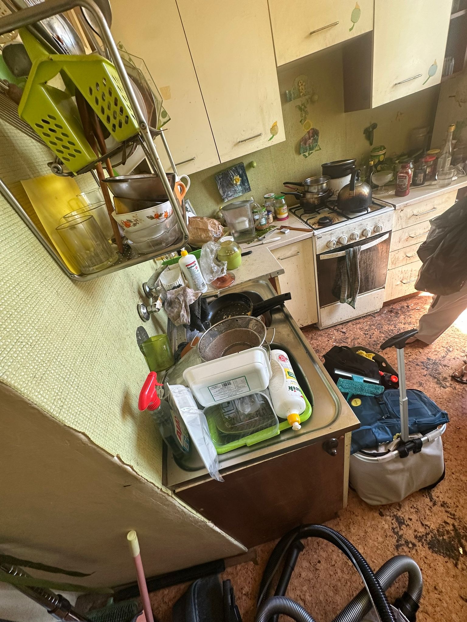 Zaniedbana kuchnia z brudnymi naczyniami w zlewie, zakurzonymi szafkami i zabrudzoną podłogą. Widoczny sprzęt do sprzątania. Zdjęcie 'przed' sprzątaniem.