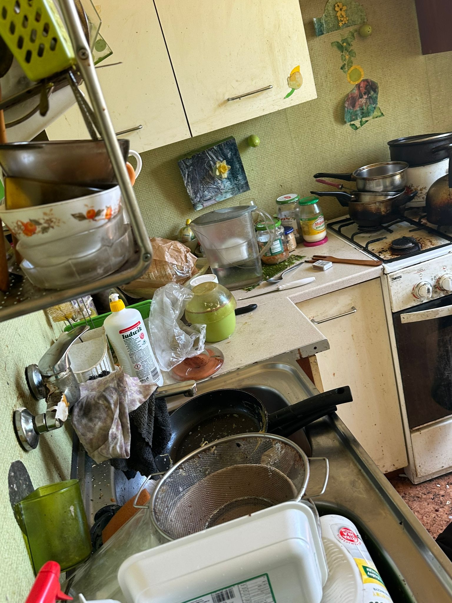 Zaniedbana kuchnia: brudny zlew pełen naczyń, tłusty piekarnik i kuchenka. Widoczne środki czystości i akcesoria kuchenne w nieładzie.