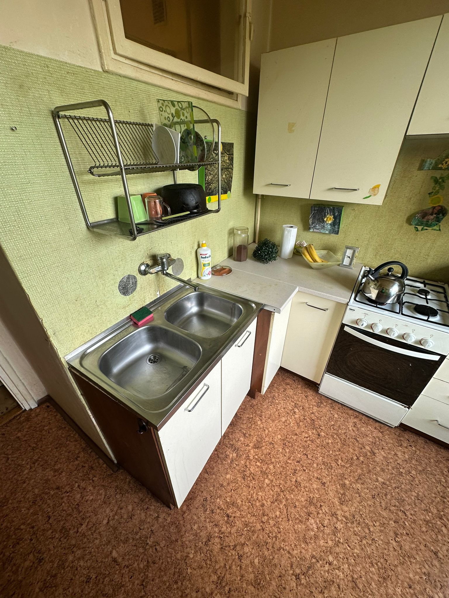 Umyta, dwukomorowa, stalowa zlewozmywak w kuchni z zielonymi ścianami. Na suszarce naczynia, obok płyn do naczyń. Na kuchence czajnik. Widok z góry.