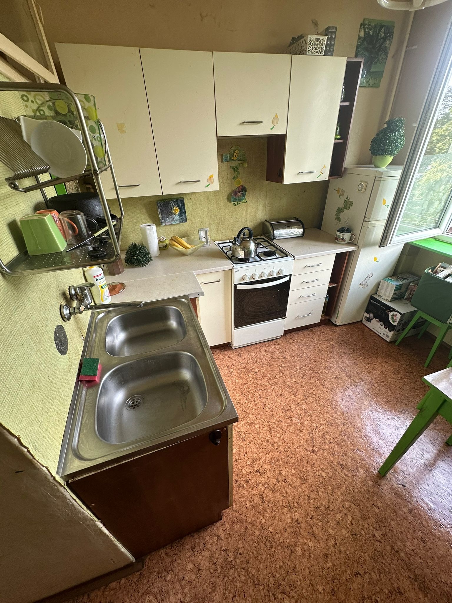 Umywalka dwukomorowa ze stali nierdzewnej w kuchni z zielonymi ścianami i beżowymi szafkami. Widok na kuchenkę i lodówkę.