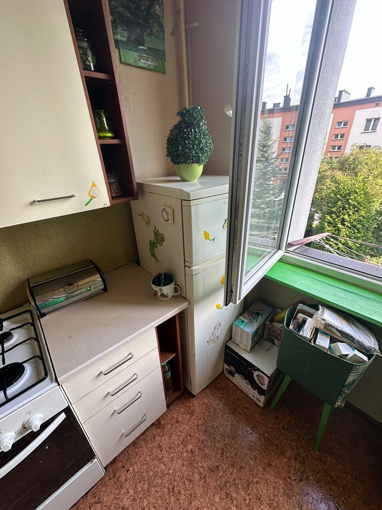 Uporządkowana kuchnia z otwartym oknem, widok na zieleń. Na blacie chlebak i kuchenka. Na szafkach naklejki. Na podłodze pudełka i kosz.