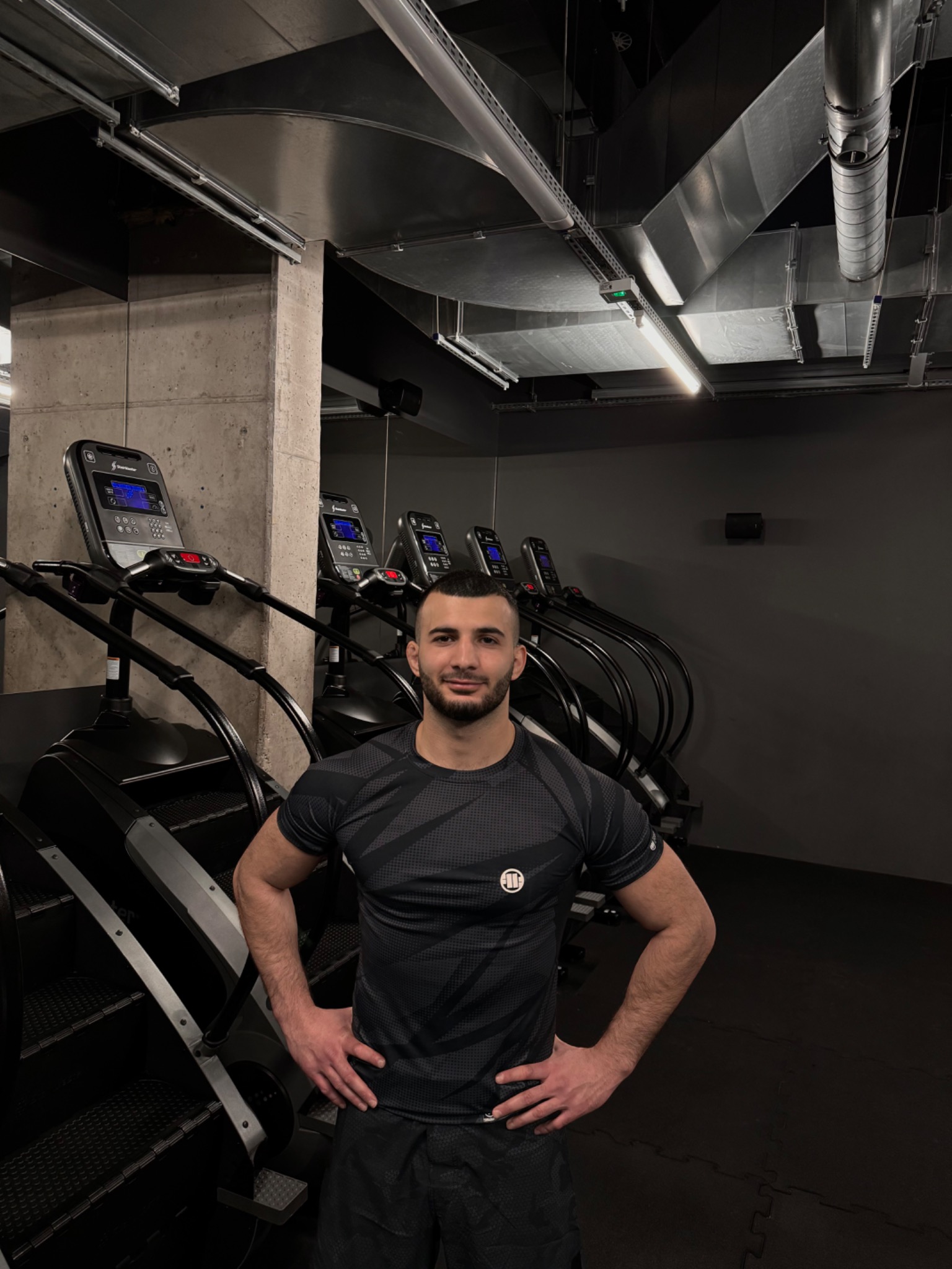 Trener personalny pozuje w siłowni obok schodów treningowych, widoczna surowa betonowa ściana i instalacja wentylacyjna pod sufitem.