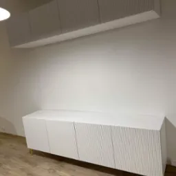 Nowoczesna, biała szafka RTV z ryflowanymi frontami i złotymi nóżkami, wisząca szafka nad nią, minimalistyczne wnętrze.