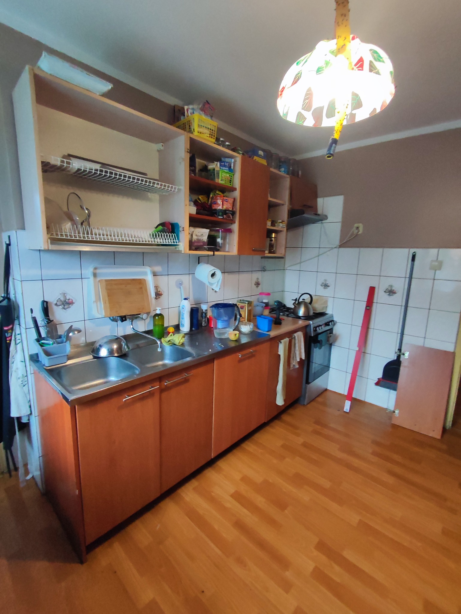 Widok na kuchnię z drewnianymi frontami szafek, dwukomorowym zlewem ze stali nierdzewnej i otwartymi szafkami wiszącymi z różnorodnymi produktami spożywczymi i akcesoriami kuchennymi, oświetloną...