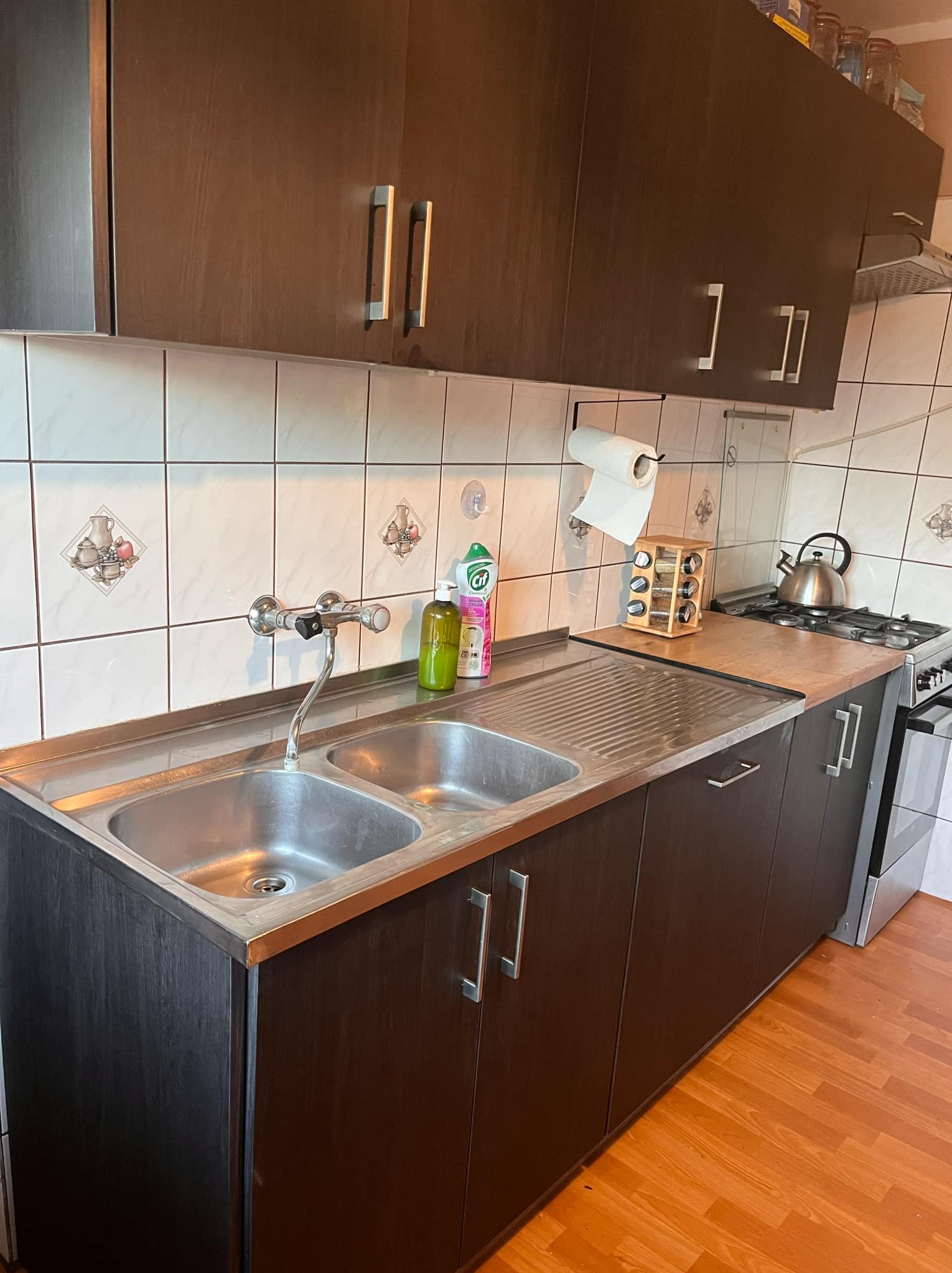 Ciemna zabudowa kuchenna z dwukomorowym zlewem ze stali nierdzewnej, bateria kuchenna, białe płytki z dekoracyjnymi motywami, przyprawy w drewnianym stojaku, czajnik na płycie gazowej.