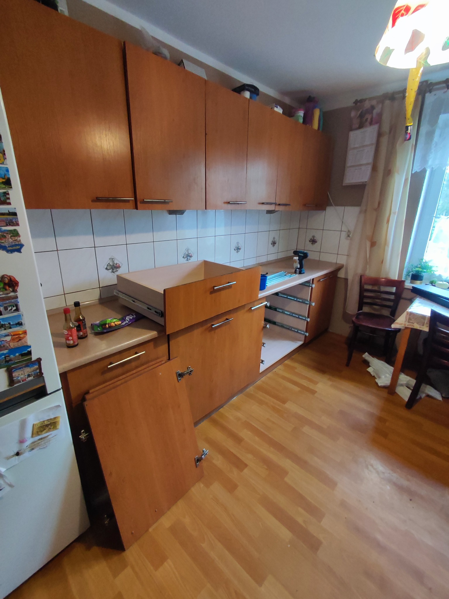 Montaż drewnianej zabudowy kuchennej: widoczna wysunięta szuflada z prowadnicami, odkręcone drzwiczki szafki oparte o białą lodówkę, w tle wkrętarka na blacie.