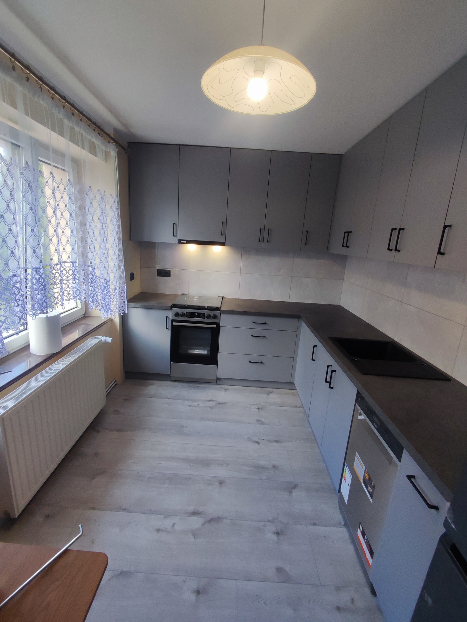 Szara zabudowa kuchenna z ciemnym blatem, widok na okno z firanką i grzejnik. Prosta lampa sufitowa.