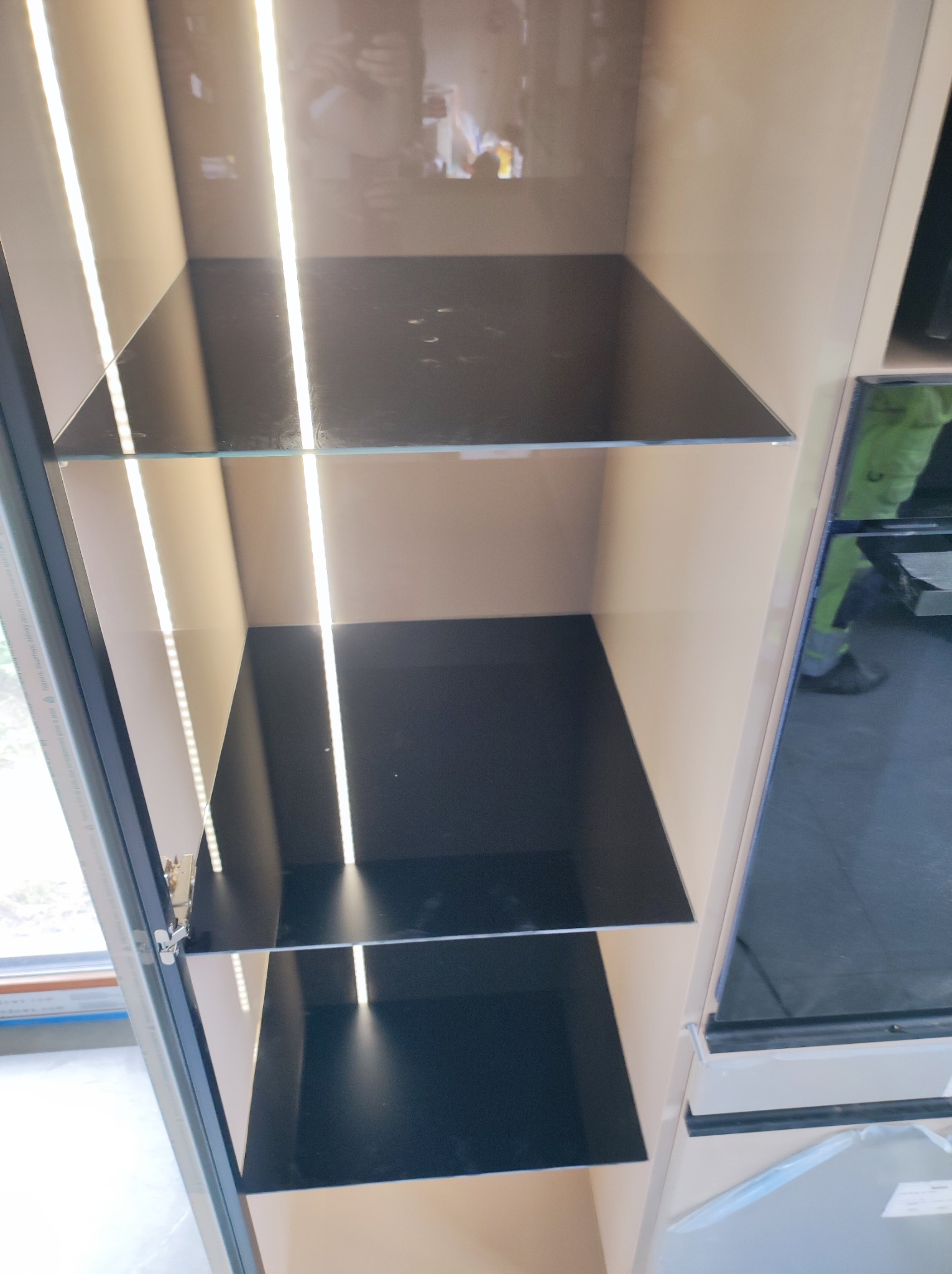 Wnętrze szafy z podświetlanymi półkami z czarnego szkła, widoczny fragment okna i odblask osoby w szybie.