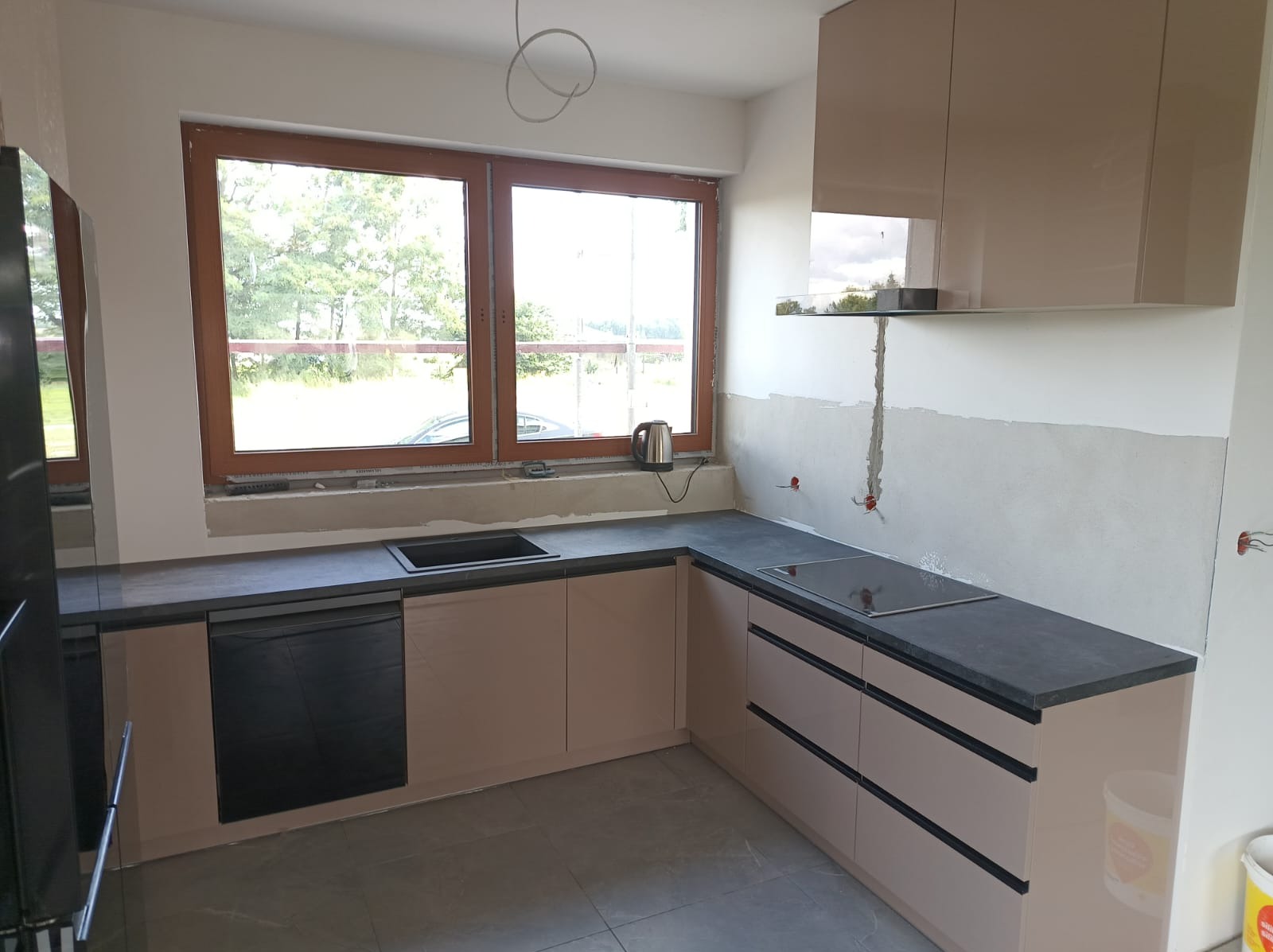 Montaż kuchni: beżowe szafki z czarnymi blatami i oknem z widokiem na zieleń, ściany w trakcie wykończenia.